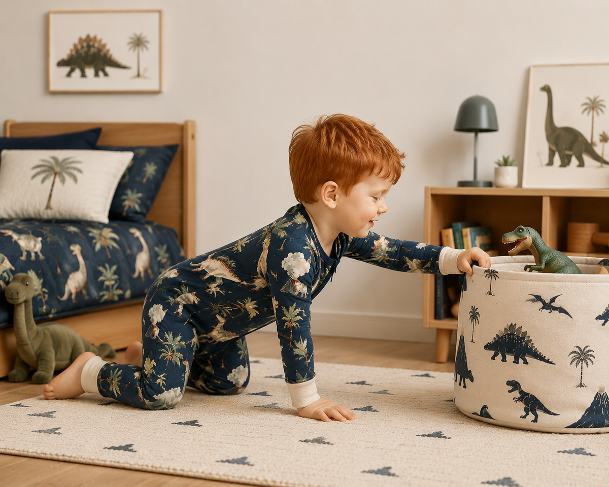 A young boy with red hair crawls on a patterned rug in a child's bedroom, wearing a navy blue dinosaur-print pajama set and reaching into a matching fabric storage bin.