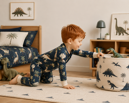 A young boy with red hair crawls on a patterned rug in a child's bedroom, wearing a navy blue dinosaur-print pajama set and reaching into a matching fabric storage bin.